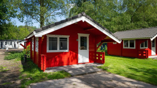 Cabaña roja con puerta y ventanas blancas, situada en una zona verde y arbolada bajo la luz del sol.