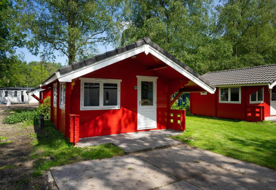 Cabaña roja con puerta y ventanas blancas, situada en una zona verde y arbolada bajo la luz del sol.