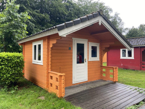 Kleine houten blokhut met veranda in Het Amsterdamse Bos, Nederland, omringd door bomen en gras.