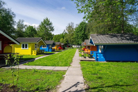 Cabañas de glamping coloridas en un camping verde con mesas, senderos y árboles bajo el sol brillante.
