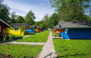 Cabañas de glamping coloridas en un camping verde con mesas, senderos y árboles bajo el sol brillante.