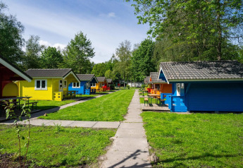 Cabine glamping colorate in un campeggio verde con tavoli da picnic, alberi e viottoli sotto il sole.