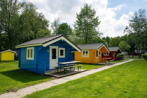 Cabañas glamping coloridas sobre césped con árboles y mesas de picnic bajo un cielo parcialmente nublado.