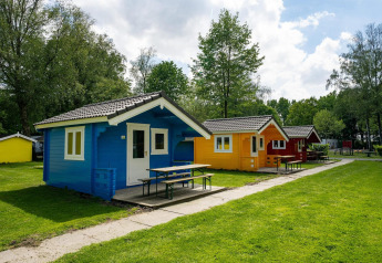 Colorate cabine glamping su prato, circondate da alberi e tavoli da picnic, sotto cielo parzialmente nuvoloso.