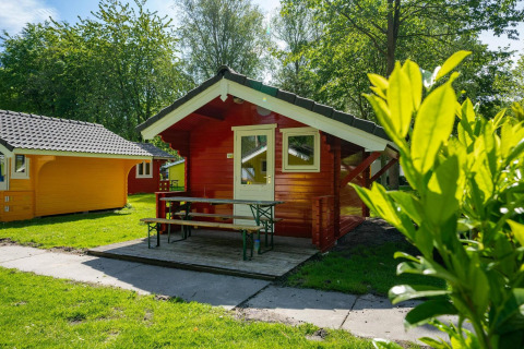 Cabañas glamping rojas y amarillas con porche y bancos, rodeadas de naturaleza verde y soleada.