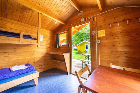Interieur van een glampinghut met houten wanden, stapelbed, tafel en uitzicht op gele hut buiten.