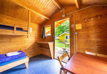Interno di una cabina glamping in legno con letti a castello, tavolo e vista su una cabina gialla all’esterno.