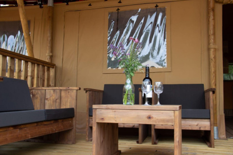Buitenzitje van een safaritent bij Het Amsterdamse Bos met houten meubels, een vaas bloemen en wijn.