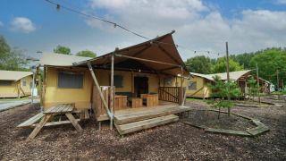 Lodge Glampingtent con grandes tiendas, terraza de madera y bancos, rodeada de árboles y luces exteriores.