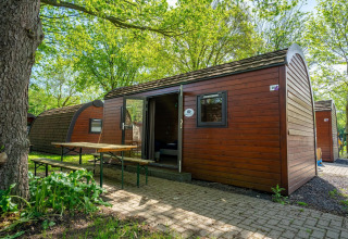 Casa pequeña llamada Pod en Het Amsterdamse Bos, Países Bajos, con bancos exteriores y vegetación abundante.