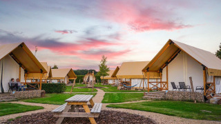Glamping site with stylish tents, picnic benches, and people relaxing at sunset under a colorful sky.