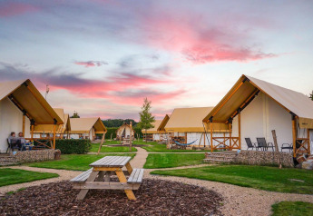 Glamping site with stylish tents, picnic benches, and people relaxing at sunset under a colorful sky.