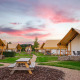 Glamping-lejrplads med luksustelte og picnicborde under en farverig solnedgangshimmel.