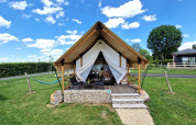 A photo of a stylish glamping tent cabin featuring a porch, outdoor seating, and green lawn under blue sky.
