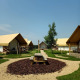 Et glamping-sted med moderne telte, træborde og grusstier under en klar blå himmel og solskin.