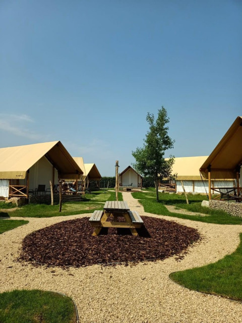 Site de glamping avec tentes modernes, table de pique-nique et allées gravillonnées sous ciel bleu.