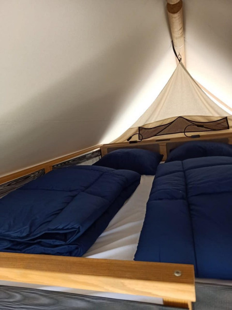 Interior de una tienda glamping con una cama doble acogedora, ropa de cama azul y almohadas sobre un marco de madera.