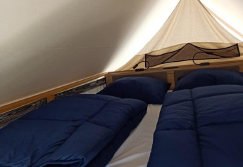 Interior de una tienda glamping con una cama doble acogedora, ropa de cama azul y almohadas sobre un marco de madera.