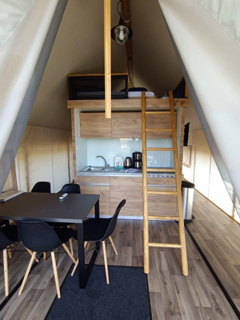 Interior de una tienda de glamping con cocina, mesa de comedor, sillas negras, piso de madera y altillo.