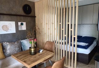 Interior moderno de una tiny house con separador de madera, área de cama, mesa comedor y sofá colorido.