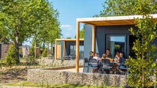 Family sitting outside Belsano lodge on a sunny day, surrounded by trees and modern vacation cabins.