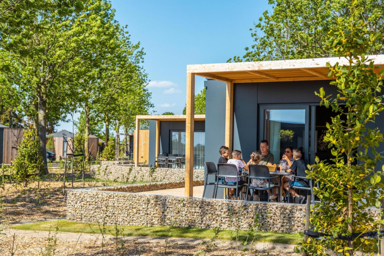 Family sitting outside Belsano lodge on a sunny day, surrounded by trees and modern vacation cabins.