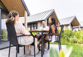 Tres personas disfrutan de una comida al aire libre en Mountain Lodge, Resort Mooi Bemelen, Países Bajos, rodeados de vegetación.
