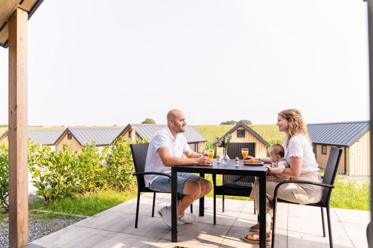 Familia desayunando al aire libre en Mountain Lodge, Resort Mooi Bemelen, Países Bajos, con cabañas de fondo.
