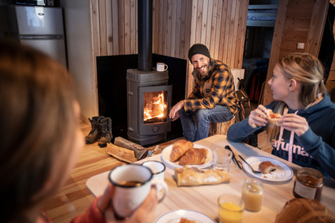 Drei Personen genießen Frühstück und Gemütlichkeit am Kamin in einer Holzhütte bei Huttopia la Clarée.