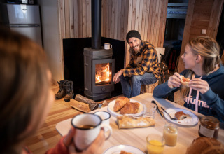 Three people share breakfast and relax by a wood stove inside a cozy cabin at Huttopia la Clarée, France.