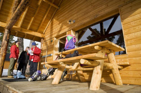 Feriepark i træhytte med folk i vintertøj, skiudstyr og bænk i Les Chalets Huttopia de Font-Romeu, Frankrig.