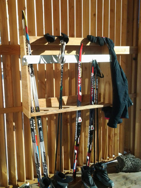 Estante para equipo de esquí con esquís, botas, bastones y chaqueta sobre una cerca de madera al sol.