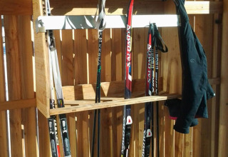 Portant à équipement de ski avec skis, bottes, bâtons et veste devant une clôture en bois ensoleillée.