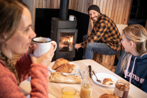 Familia desayunando croissants y bebidas calientes junto a una estufa de leña en una acogedora cabaña.