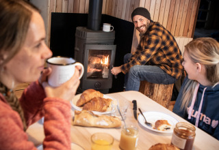 Familie frühstückt mit Croissants und Heißgetränken vor einem Kaminofen in einer gemütlichen Hütte.