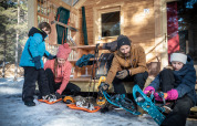 Familie legt Schneeschuhe an vor Berghütte in Les Chalets Huttopia de Vallouise, Provence-Alpes-Côte d’Azur, Frankreich.
