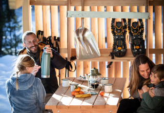 A family enjoys warm drinks and time together inside a cozy wooden cabin with snowshoes hanging behind.