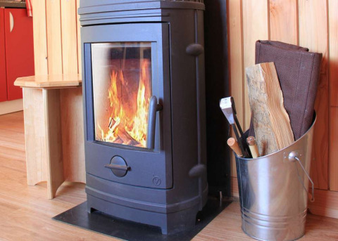 Estufa de leña encendida en una cabaña de madera, junto a un cubo metálico con leña y utensilios de chimenea.