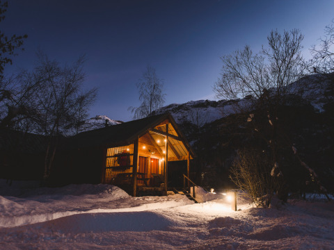 Cozy lit wooden chalet in the snow at night, nestled in mountains at Les Chalets Huttopia de Vallouise, France.