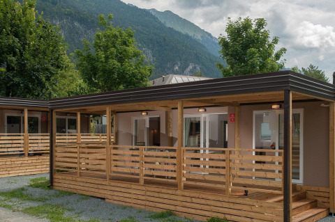 Cabaña moderna con terraza de madera, rodeada de árboles y montañas al fondo, ambiente tranquilo.