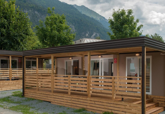 Modern vakantiehuisje met houten veranda, omgeven door bomen en bergen op de achtergrond.