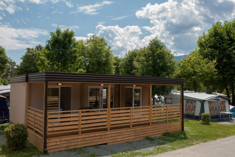 Moderne hytte ved Hermagor-Nassfeld i Østrig med træterrasse omgivet af grønne træer under en blå himmel.