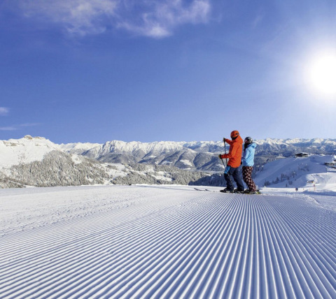Dos personas con esquís en una pista recién preparada, montañas nevadas y cielo soleado al fondo.