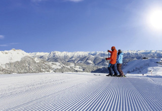 Due persone sugli sci su una pista battuta, circondate da montagne innevate e sole splendente.