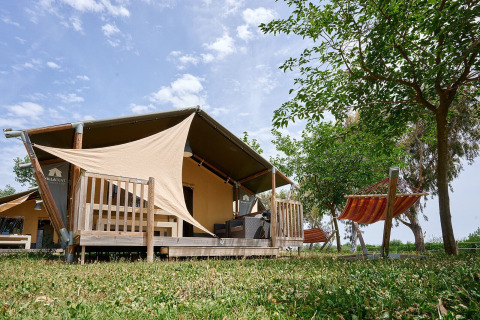 Tienda safari Villatent Wood en Camping Nautic Almata, España, con terraza de madera y hamaca en el jardín.