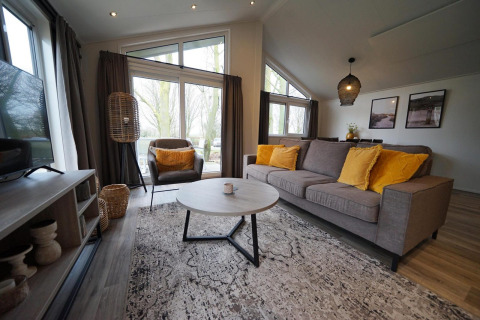 Modern living room in Hackfort lodge at Vakantiepark Aan de Maas, Netherlands, with yellow pillows and large windows.