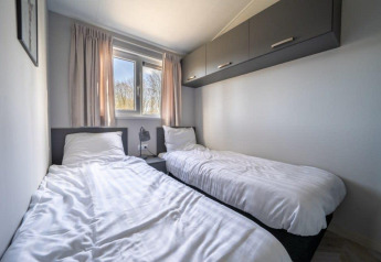 Modern lodge bedroom with two single beds, white bedding, window, and overhead cabinets at Vakantiepark Aan de Maas.