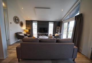 Moderne woonkamer in lodge Hackfort op Vakantiepark Aan de Maas, Nederland, met bank en grote ramen.