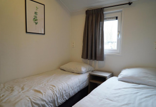 Twee eenpersoonsbedden en een nachtkastje onder een raam bij Hackfort lodge, Vakantiepark Aan de Maas.