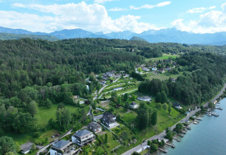 Vista aérea de un alojamiento glamping junto al lago, rodeado de colinas verdes y montañas al fondo.